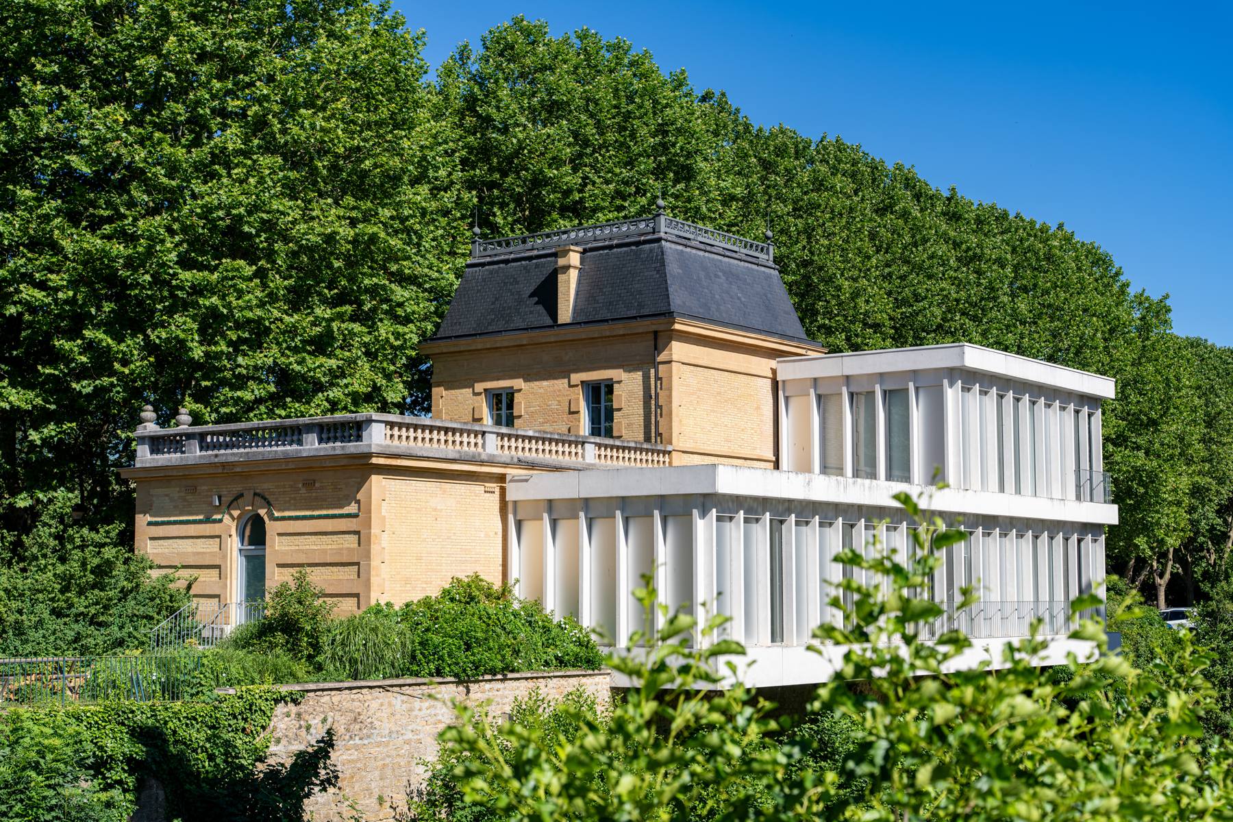 Pavillon de pierre prolongé par une extension vitrée blanche au milieu des arbres, Frechin